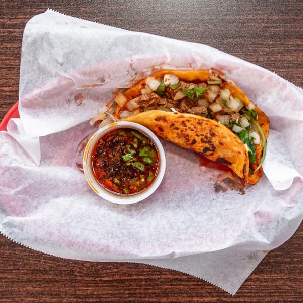 Two birria tacos with meat, onions, and cilantro, served with a side of consommé.