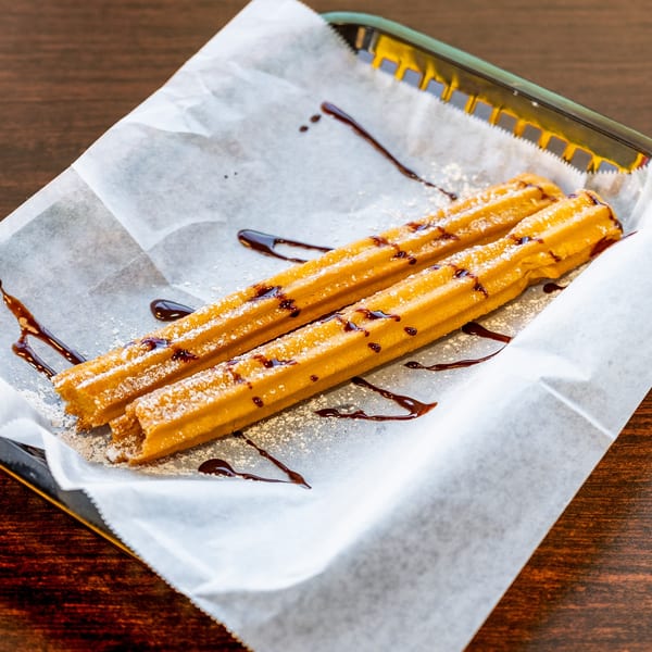 Two churros dusted with powdered sugar and drizzled with chocolate sauce in a basket.