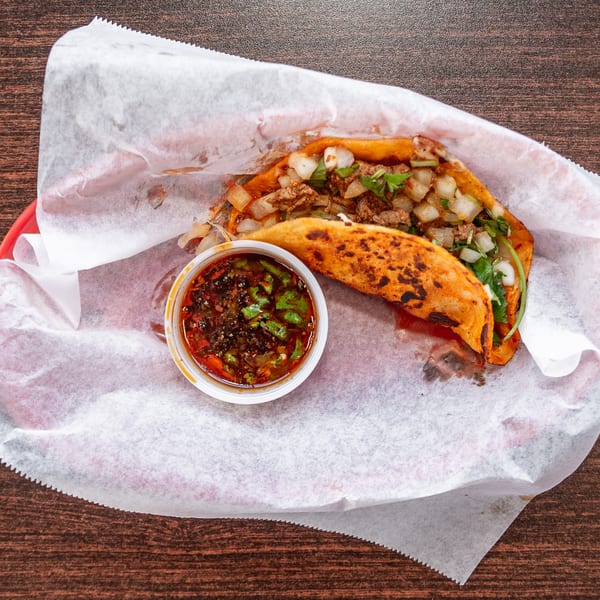 Two birria tacos with diced onions, cilantro, and a side of consommé on a wooden table.