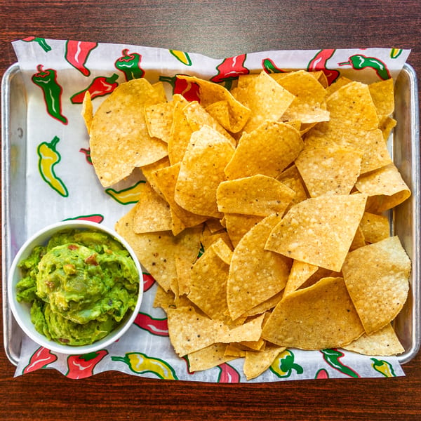 Uno Dos Tacos' tortilla chips and guacamole in a metal tray with chili pepper-patterned paper.