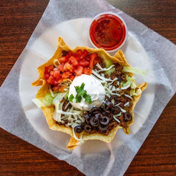 A taco salad in a crispy tortilla bowl with ground beef, lettuce, tomatoes, olives, cheese, and sour cream.